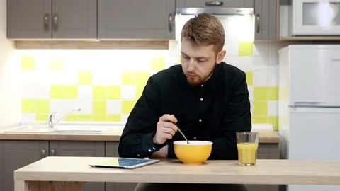 Young man with tablet eating breakfast sitting by table in kitchen at home. Stockbeeldmateriaal 103940290