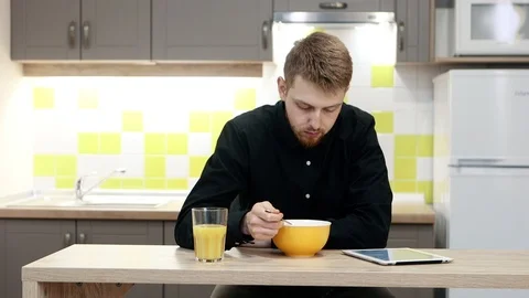 Young man with tablet eating breakfast sitting by table in kitchen at home. Stockbeeldmateriaal 104668924