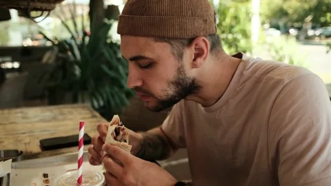 A young man takes a bite of a burrito while sitting in an outdoor cafe on a Stock Footage 342545295