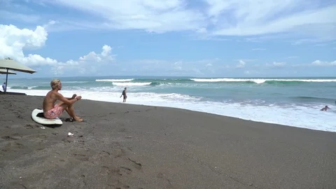 Young man takes a break from surfing on a dark sand beach Video stock 74288513