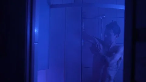 Young man taking a shower behind a foggy glass door, illuminated by moody blue Stock Footage 305446811