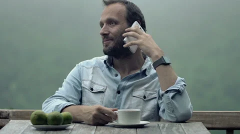 Young man talking on cellphone while drinking coffee by table on terrace HD Stock-Footage 55679462