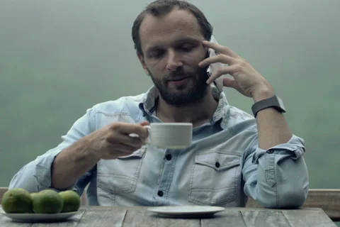 Young man talking on cellphone while drinking coffee by table on terrace NTSC Stock Footage 55679485