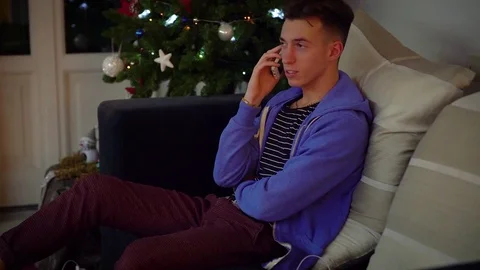 A young man talking on his cell phone  with a Christmas tree in the back grou 스톡 동영상 80023329