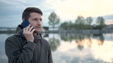 Young man talking on phone in the forest near lake. He has a vacation on nature. Stock Footage 99433283