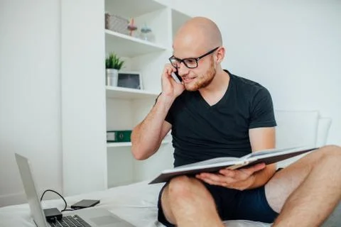 Young man is talking by the phone, using laptop pc and holding notepad. Home  Stock Photos