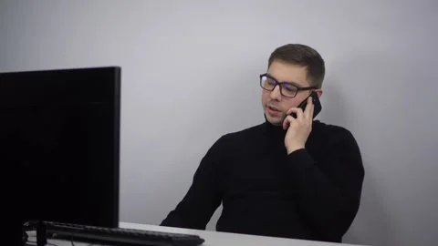 A young man is talking on the phone while sitting at a computer in the office Stock Footage 231059278