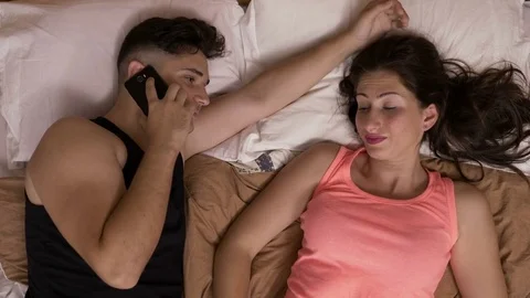 Young man talking at smartphone in bed beside his girlfriend caressing her ha Stock Footage 80177236