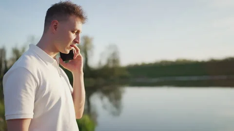 Young Man Talking on Smartphone Outdoors by Lake at Sunset Stock Footage 339031383