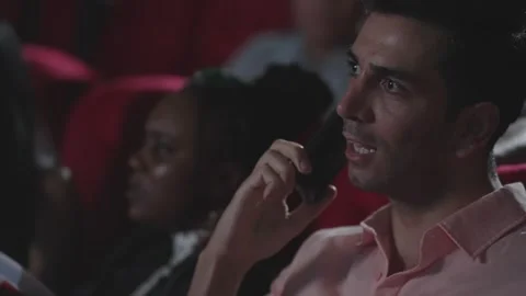 Young man talking on smartphone watching movie at cinema and disturbing audience Stock Footage 248599619