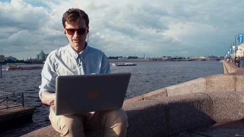 Young man talking on video calling with laptop sitting on bench near seafront Stock Footage 77167475