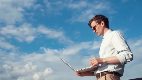Young man talking on video calling with laptop standing outdoors. Summer day Stock Footage 77167737