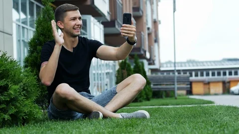 Young man talking in video chat via cell phone, sitting on the grass and speak Stock Footage 110755691