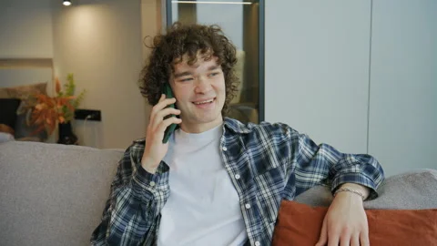 Young man talks on the phone while sitting on a couch in a modern room during th Stock Footage 345299956