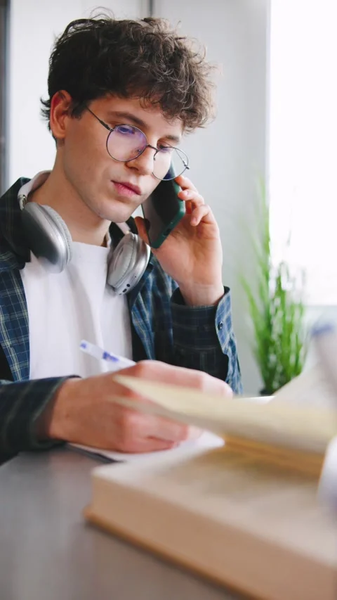Young man talks on the phone while writing notes at a desk in a bright room with Video stock 346186333