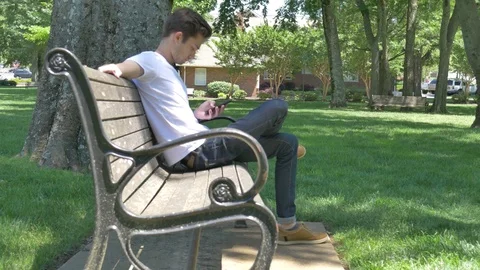 Young man texting on a bench in a park outside 스톡 동영상 75659042