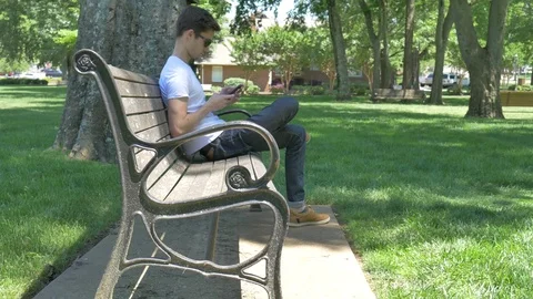 Young man texting on a bench in a park outside 스톡 동영상 75676095