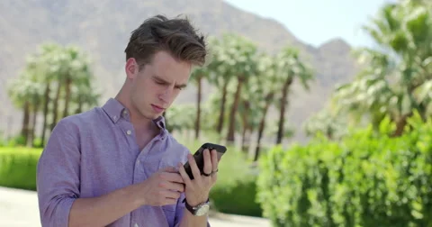 Young man texting on phone in front of palm trees and mountains 4K Stock Footage 63870861