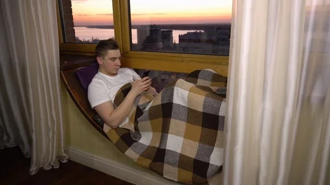A young man is texting on the phone. A man lies on a window sill by the window 库存影片 126390323
