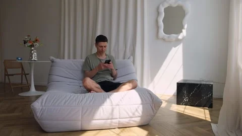 Young man texting on phone while sitting on couch in cozy modern apartment. Stock Footage 311763180