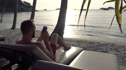 Young man texting on smartphone lying on sunbed on the beach, 4K Stock Footage 59133527