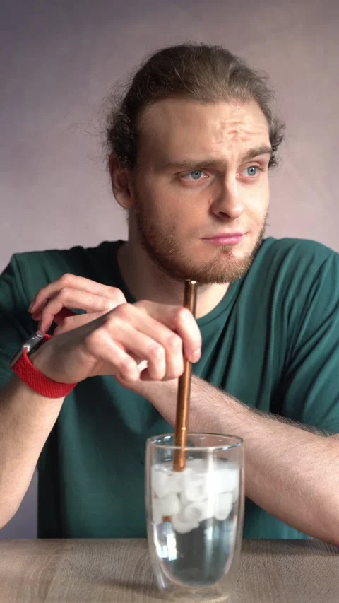 Young man thoughtfully stirring ice water with metal straw Stock Footage 229651358