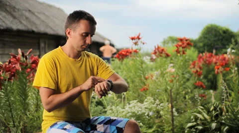 Young man touching smart watch near flowers in the garden. Checks messages Stock Footage 65093614