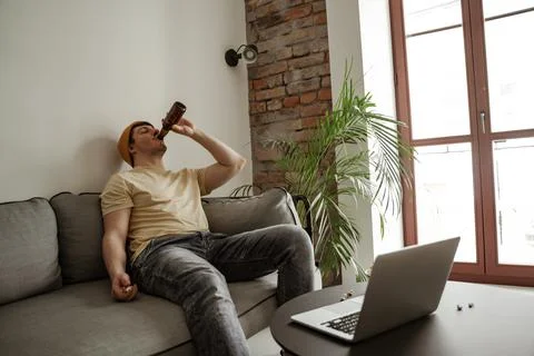 Young man tourist in yellow hat drinking beer with laptop in hotel room, male on Stock Photos