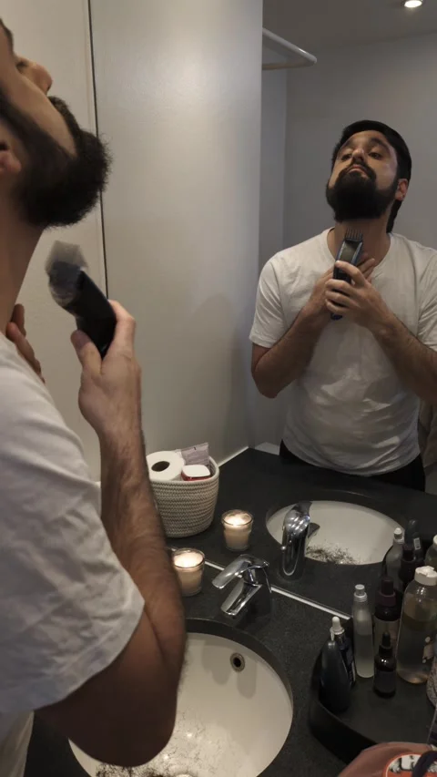 Young man trimming beard with electric trimmer in bathroom mirror Video stock 335636300