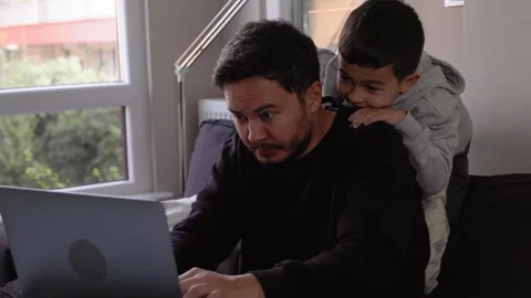 A young man trying to work at home with little son jumping on a couch. Remote Stock Footage 245799751
