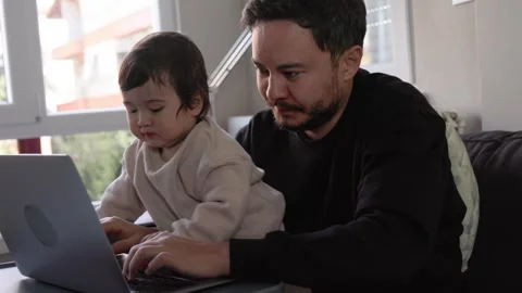 A young man trying to work on laptop holding his son at home. Remote work Stock Footage 245424702