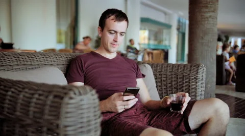 Young man type on cellphone and drinking whiskey while sitting on sofa at hotel Video stock 68642383