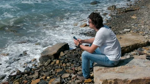 Young man types a message on mobile phone sitting on the big stone on sea beach. Stock Footage 116022242