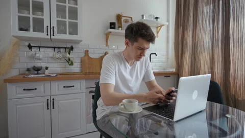 Young man types message on phone sitting by laptop at table Stock Footage 201184750