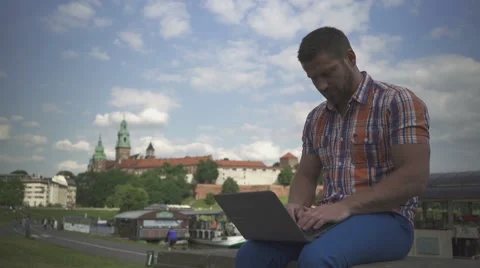 Young man typing on computer, sitting on wall by river. 库存影片 52696170