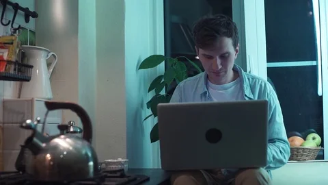 Young man typing on computer sitting at kitchen while kettle boiling on stove Stock Footage 83871347