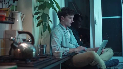 Young man typing on computer sitting at kitchen while teakettle boiling on stove Stock Footage 83871730