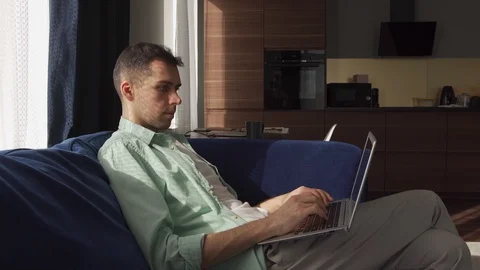 Young man typing on laptop sitting on sofa at home Stock Footage 128808089