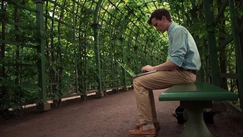 Young man typing message at laptop sitting on bench at city park. Summer day Stock Footage 77169112