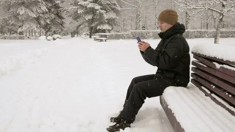 Young man typing a sms message on the phone in the winter snowy park. He smiles Stock Footage 71219348
