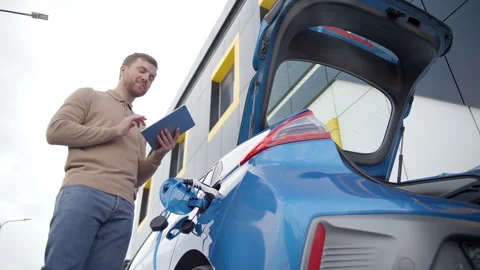 Young man typing on tablet device while waiting till his electric car charge 動画素材 257809890