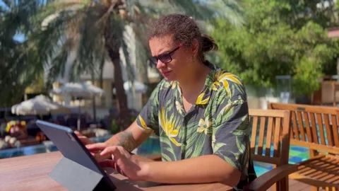 A young man typing on a tablet screen while sitting by the pool in beachwear Stock Footage 288579528