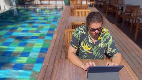 A young man typing on a tablet screen while sitting by the pool in beachwear Video stock 288579533