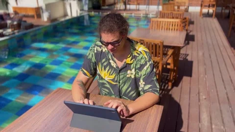 A young man typing on a tablet screen while sitting by the pool in beachwear Video stock 288579551