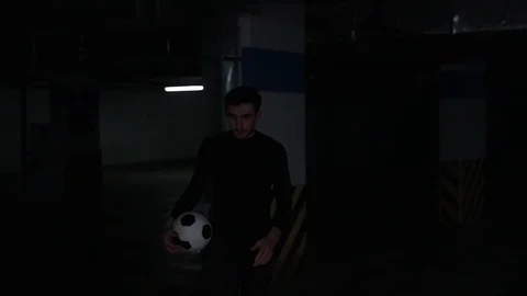 A young man on the underground parking coming from the dark holding a football Stock Footage 105562557