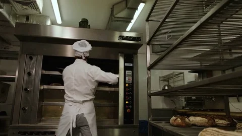 Young man in the uniform removing breads from oven and putting it to the reck Stock Footage