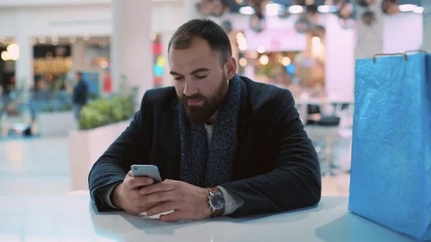 Young man use phone sitting look at camera smiling shrugs in mall feel happy Vidéo 84607895