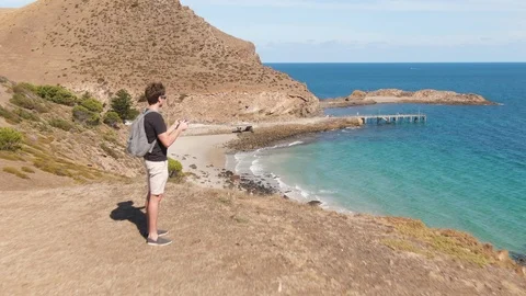 A young man uses a drone while standing on top of a hill near pristine waters in 스톡 동영상 104583732
