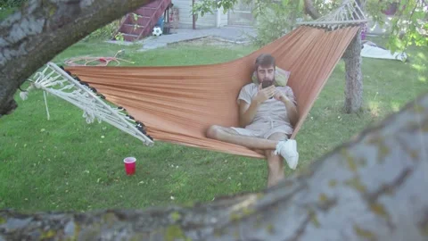 A young man uses phone while lying in a hammock in the back yard Video stock 143486153