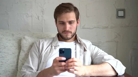 Young Man Uses Smartphone while Sitting on a Sofa, Browses Through Internet Stock Footage 154690787
